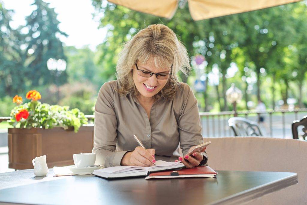 Business beautiful woman writing in business notebook using smartphone, smiling female sitting in summer outdoor cafe, coffee break