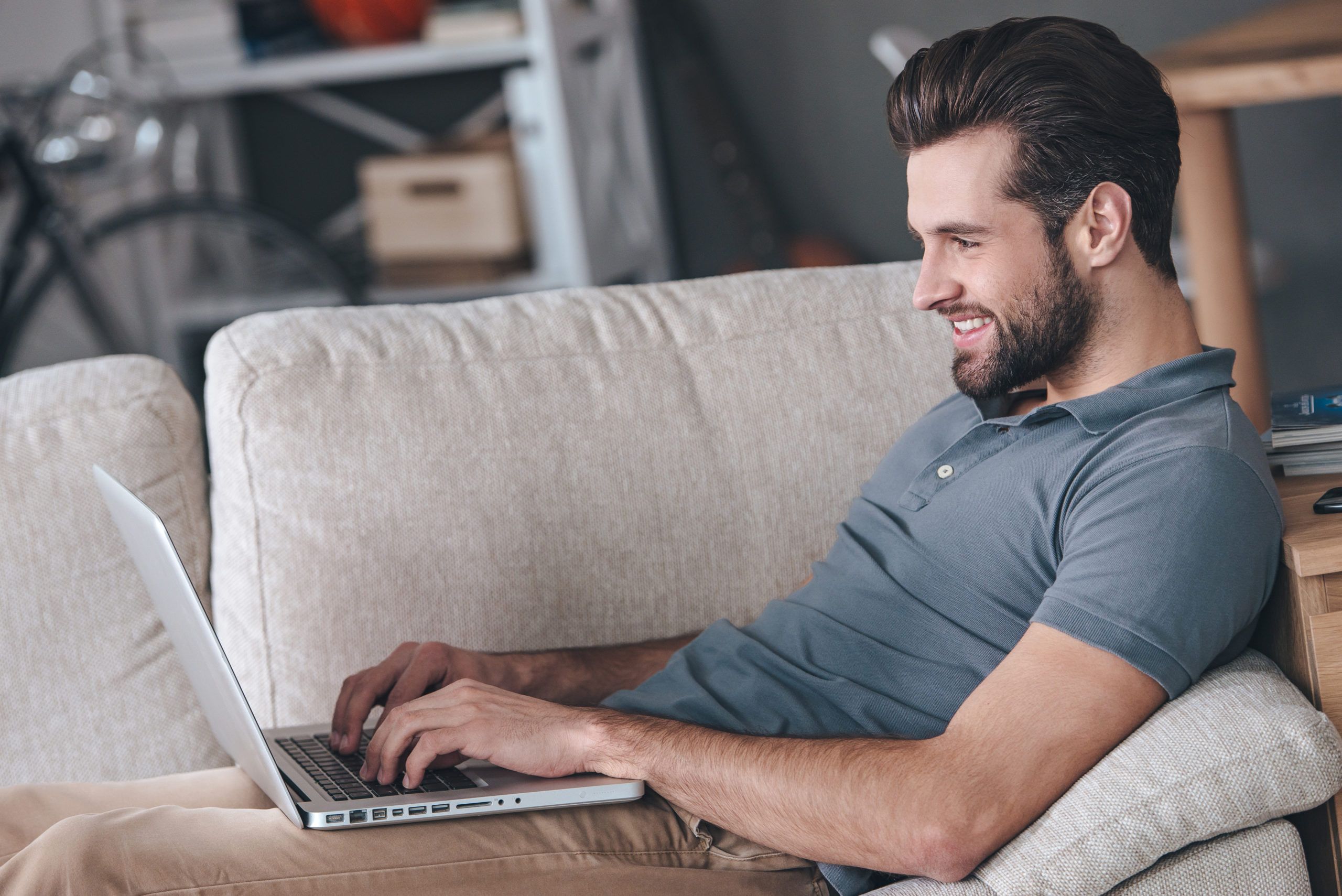 Typing new blog post. Side view of handsome young man using his laptop with smile while sitting on the couch at home
