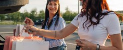 Cheerful trendy female buyers putting shopping bags into a car trunk.