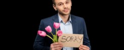 Handsome young man with pink tulips and sorry sign hopefully looking at camera isolated on black