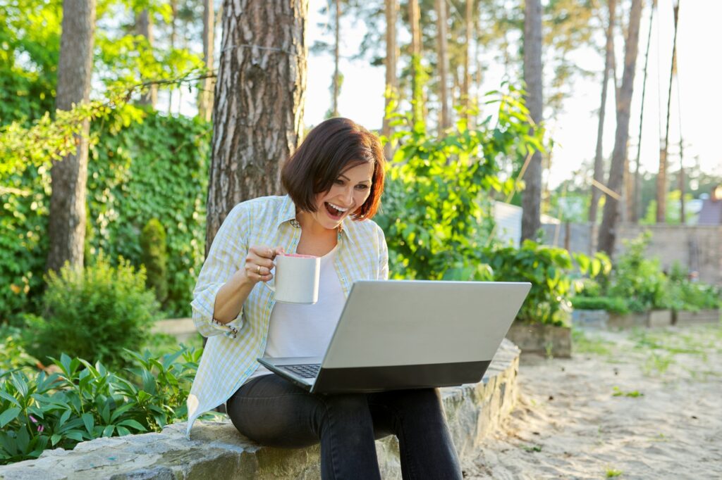 Beautiful emotional middle aged woman relaxing in garden with cup of tea and laptop