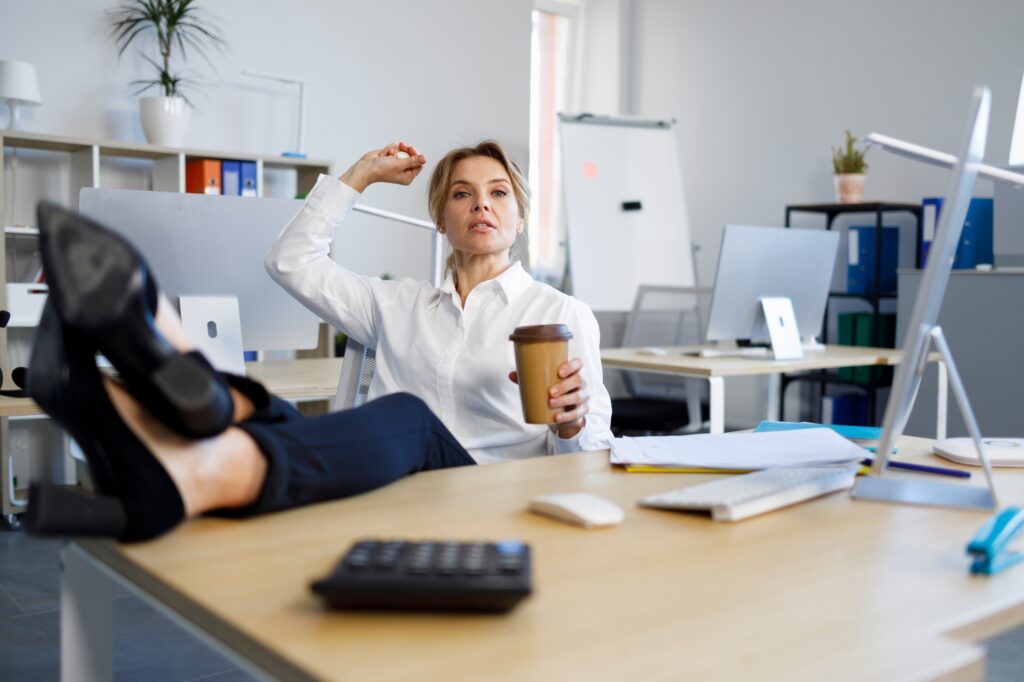Rude female boss with a cup of coffee put her legs on the desk and throws crumpled paper