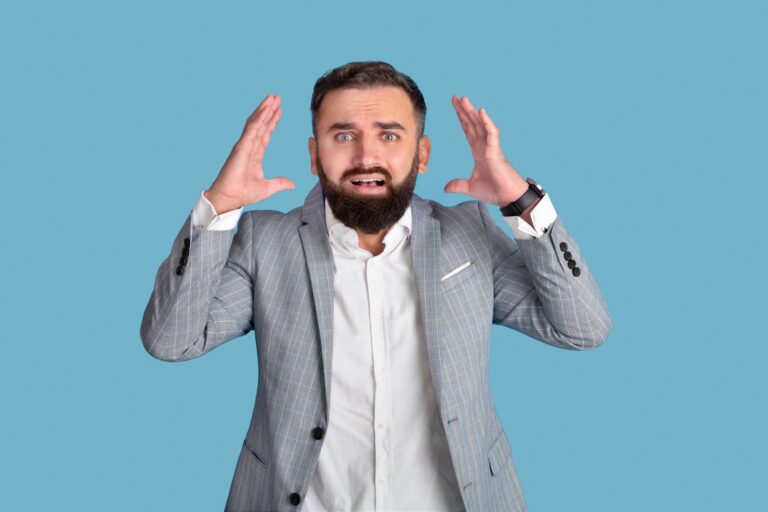 Scared young businessman grasping his head in terror on blue studio background