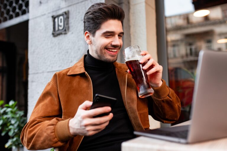 Alkoholprobléma a munkahelyen: emberi megközelítés Young attractive casual man with cellphone happily drinking beer working on laptop in cafe on street