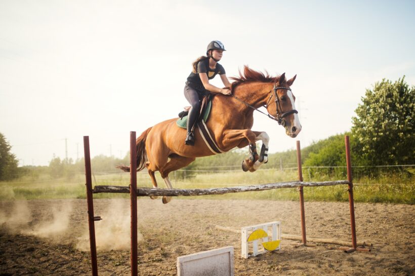 Young female jockey on horse leaping over hurdle
