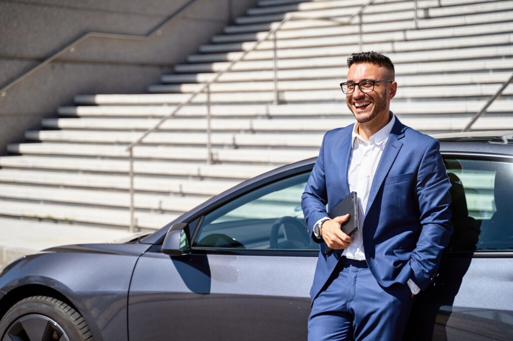 Smiling executive leaning on car, holding tablet.